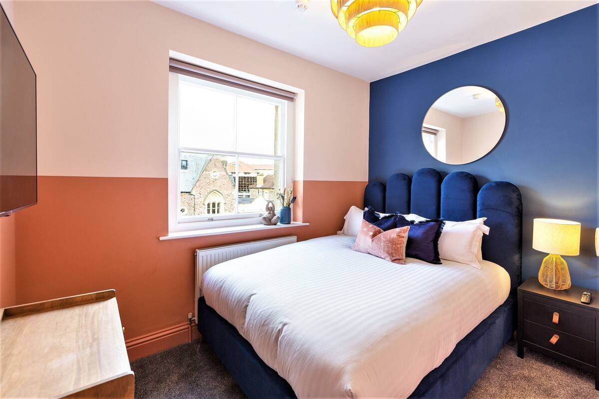 A bedroom with a large bed, a window with a view of a building, a round mirror, and a bedside lamp.