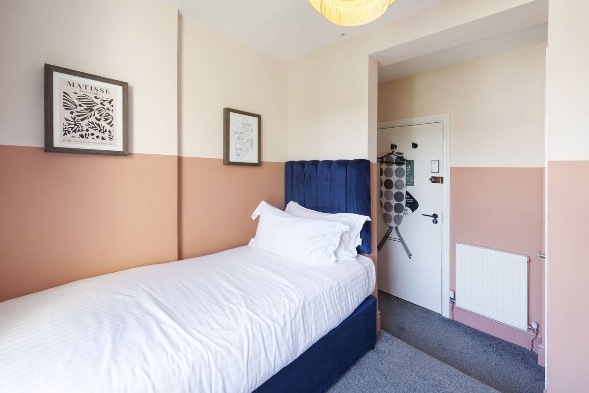 A neatly made bed with white linens and a blue headboard, a framed artwork on the wall, and a door with a radiator and a lock.