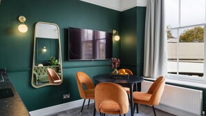 Une pièce aux murs de couleur turquoise, avec une table ronde, quatre chaises orange, un téléviseur à écran plat et une fenêtre avec des rideaux.
