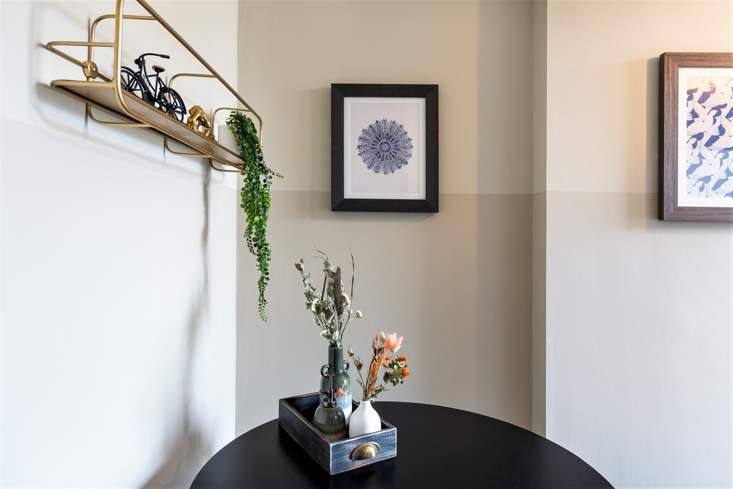 Une pièce avec une table ronde, une étagère en métal où sont posés des vélos, une œuvre d’art abstraite encadrée et un vase rempli de fleurs.