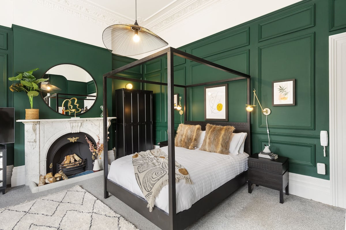 A four-poster bed with a white bedspread and decorative throw, a black nightstand with a lamp, a fireplace with logs, and a mirror on the wall.