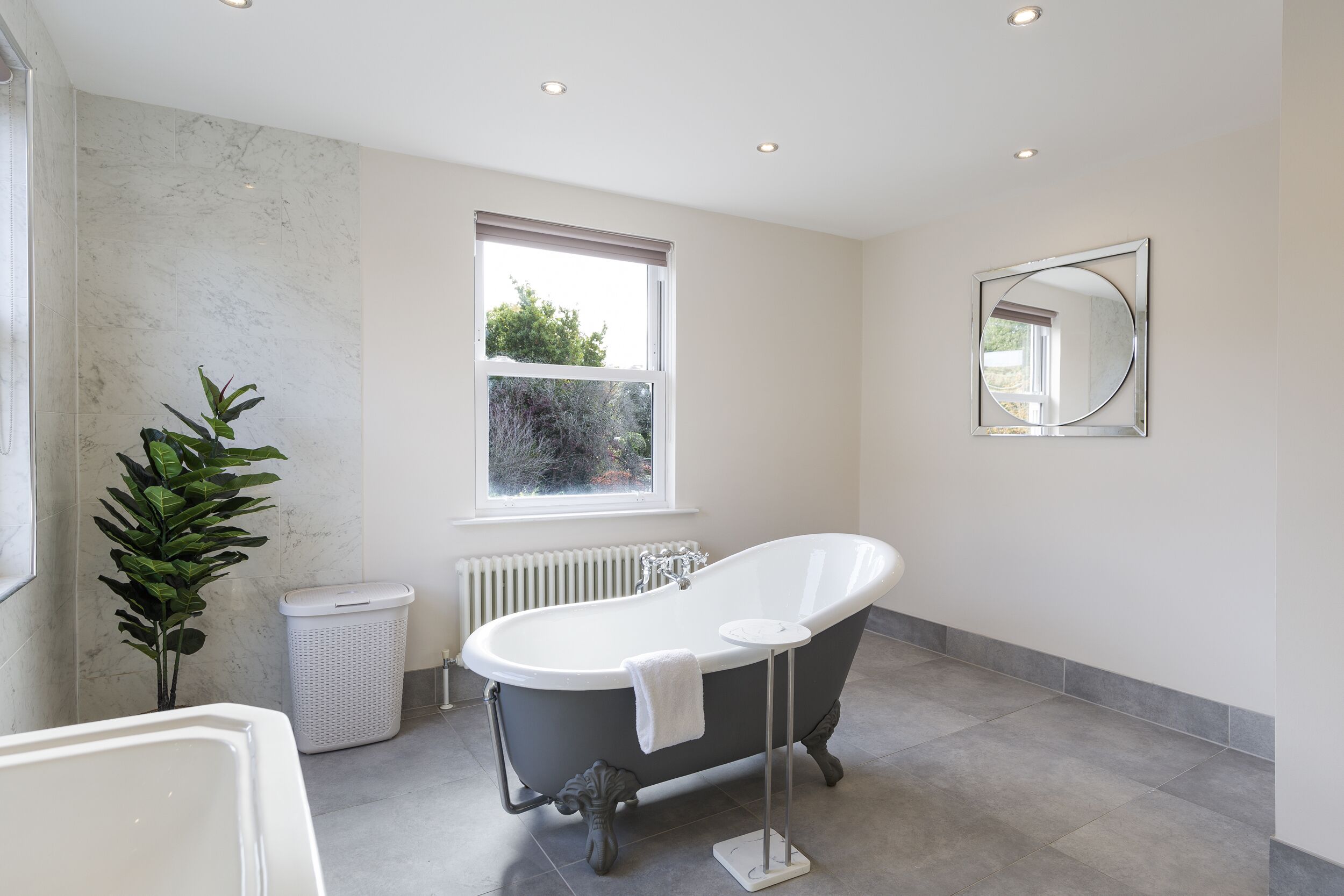 A modern bathroom with a freestanding bathtub, a window with a view, a radiator, and a potted plant.