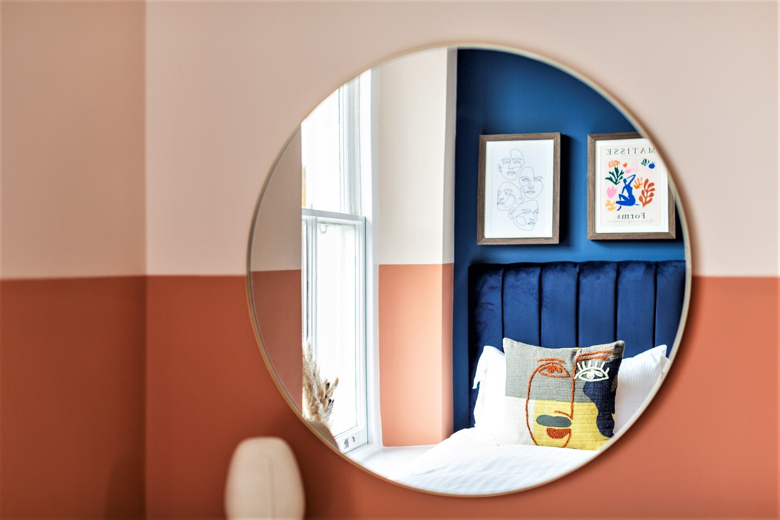 A bedroom with a round mirror reflecting the room's interior, a bed with a blue headboard, and two framed artworks on the wall.