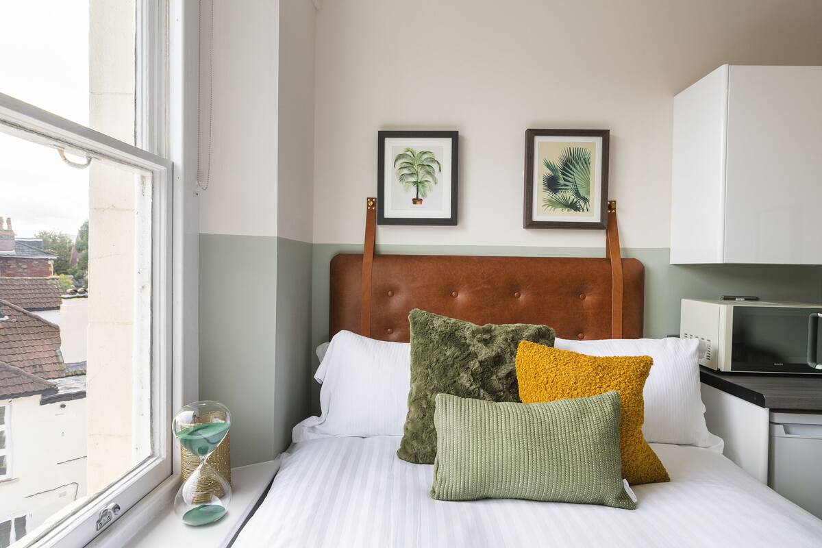 A neatly made bed with a wooden headboard, a window with a view of buildings, and a microwave on the countertop.