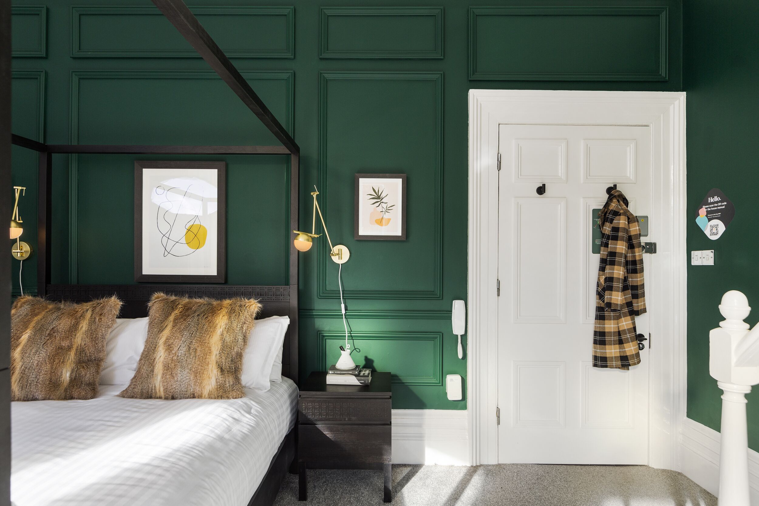 Ein Schlafzimmer mit Himmelbett, Nachttisch mit Lampe, gerahmten Kunstwerken und einem Mantel, der an der Tür hängt.