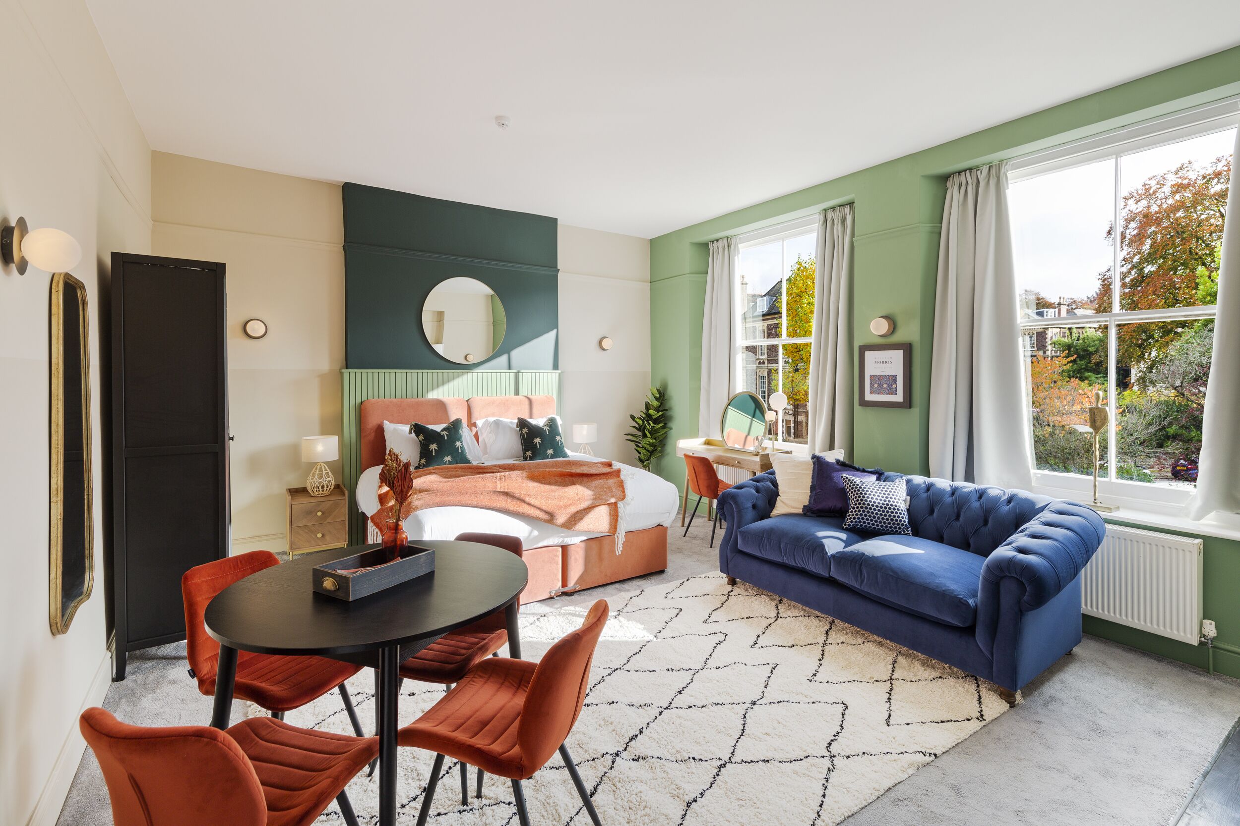 Een moderne kamer met een bed, een blauwe bank, een eettafel met stoelen en een groot raam met gordijnen.