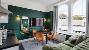 Une pièce moderne avec un mur vert, une table à manger avec des chaises, un canapé, un téléviseur et une fenêtre donnant sur des immeubles.
