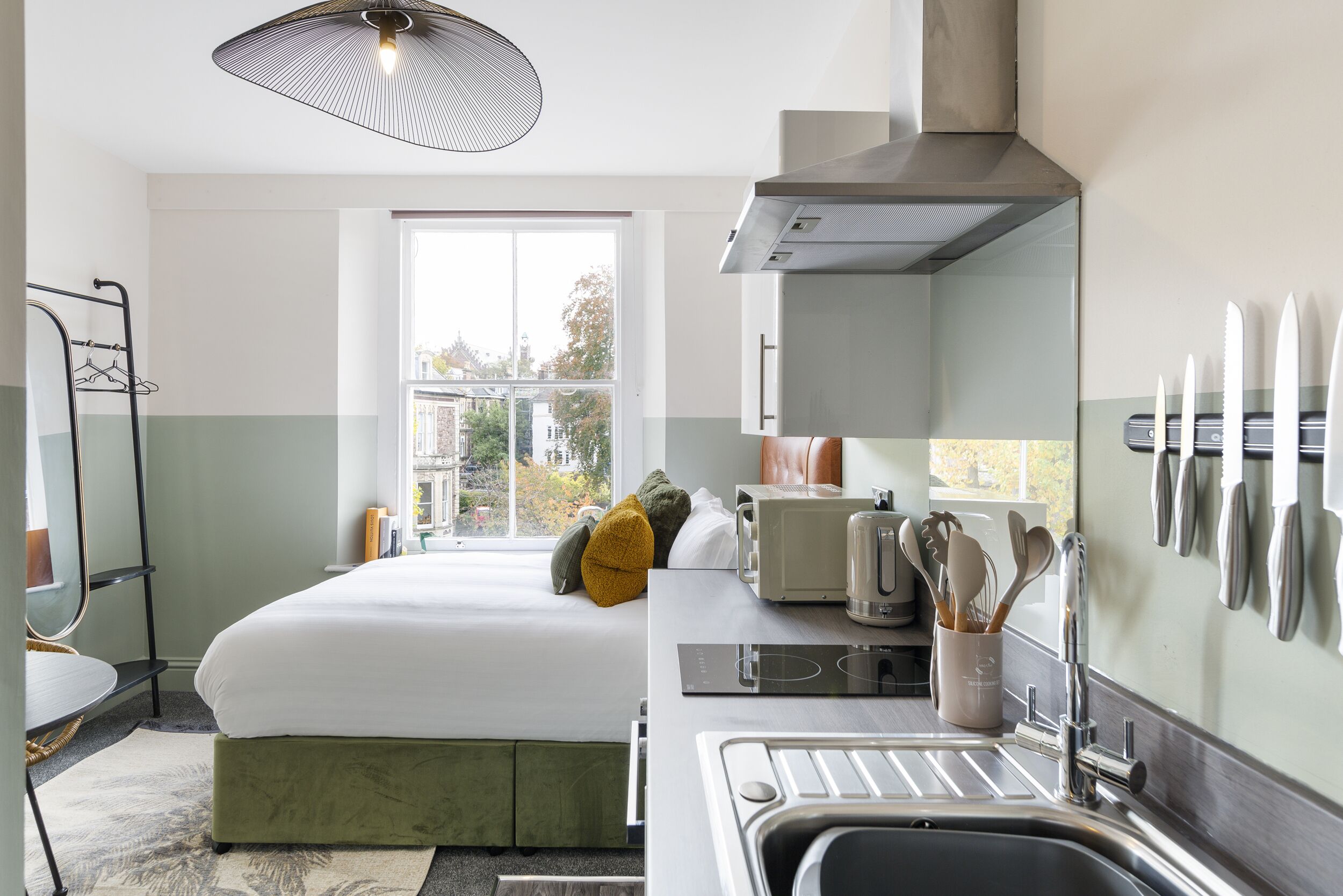 Une chambre d’hôtel moderne équipée d’un lit, d’une kitchenette et d’une grande fenêtre donnant sur la ville.