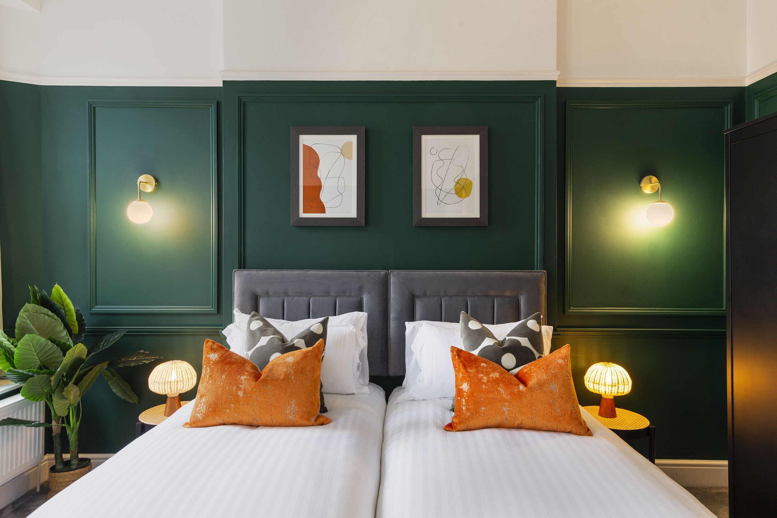 A double bed with white linens and two orange pillows, flanked by two wall-mounted light fixtures and two framed abstract artworks.