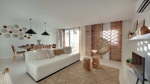 Ein modernes Wohnzimmer mit einem weißen Sofa, Holzstühlen, einem Esstisch und einem großen Fenster mit gemusterten Vorhängen.