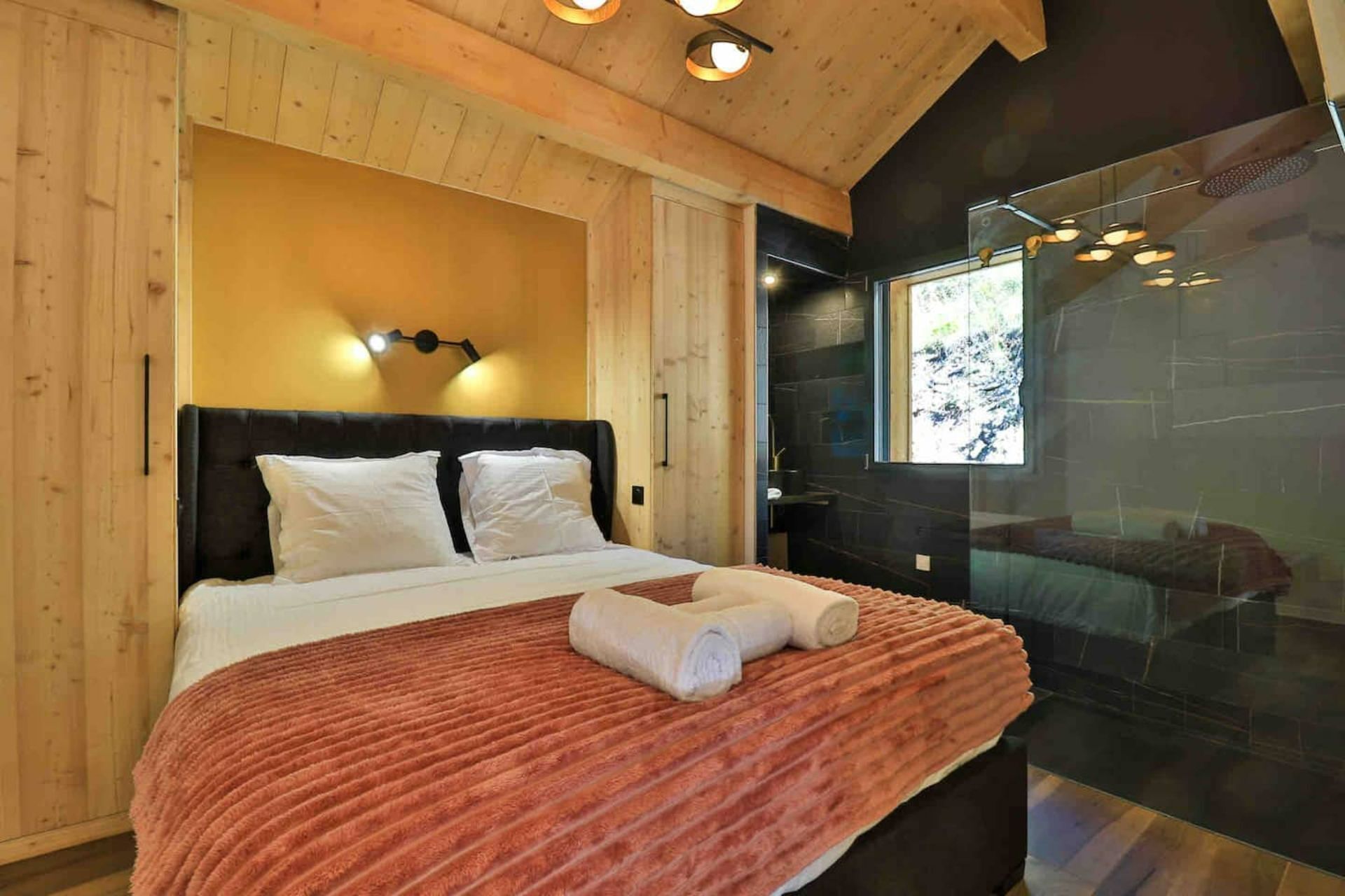 A bedroom with a wooden ceiling, a bed with white bedding, a red blanket, and a glass shower area.