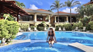 A pool area with a woman sitting by the pool, surrounded by palm trees and lounge chairs. - Stay in the best located Vacation Condo in Jaco! (Jacó)