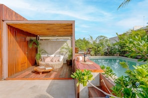 Un espacio exterior de madera con piscina, muebles de mimbre y plantas en macetas.