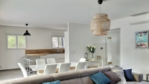 A modern living room with a sectional sofa, a dining area with white chairs, and a kitchen in the background.