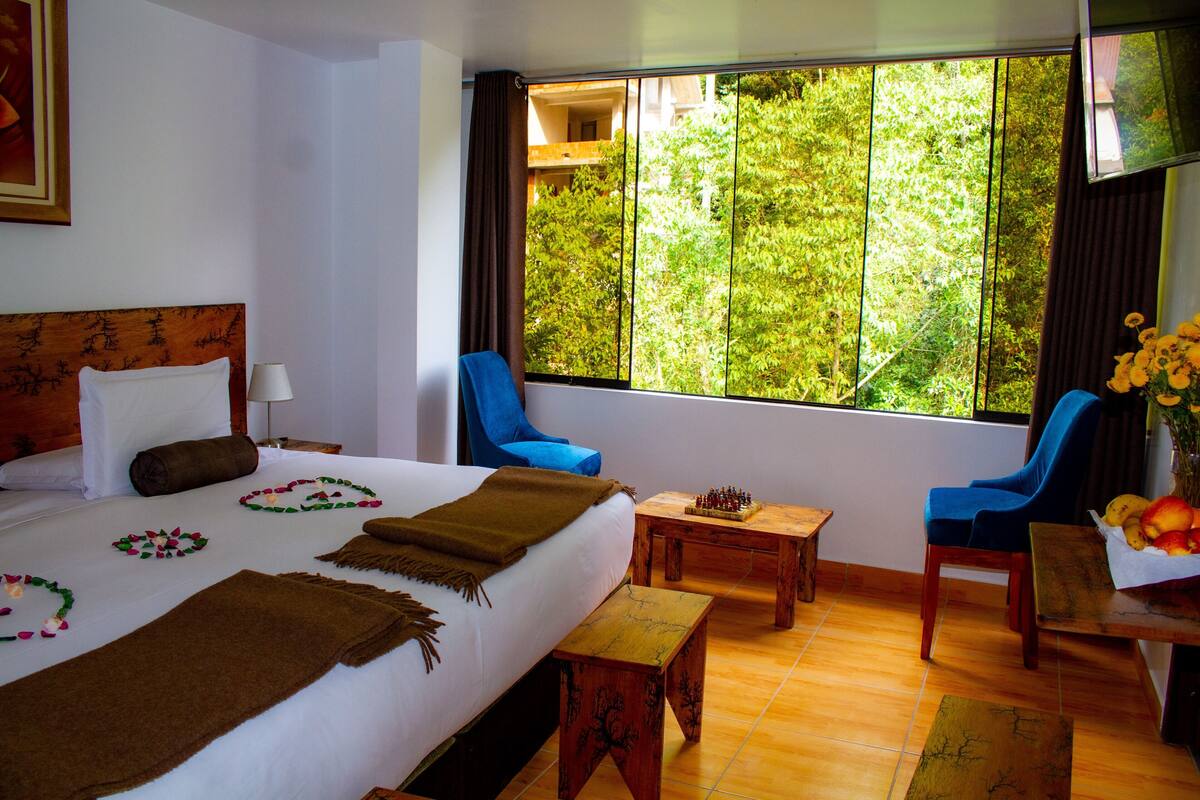 A hotel room with a bed, two chairs, a small table, and a view of greenery through the window.