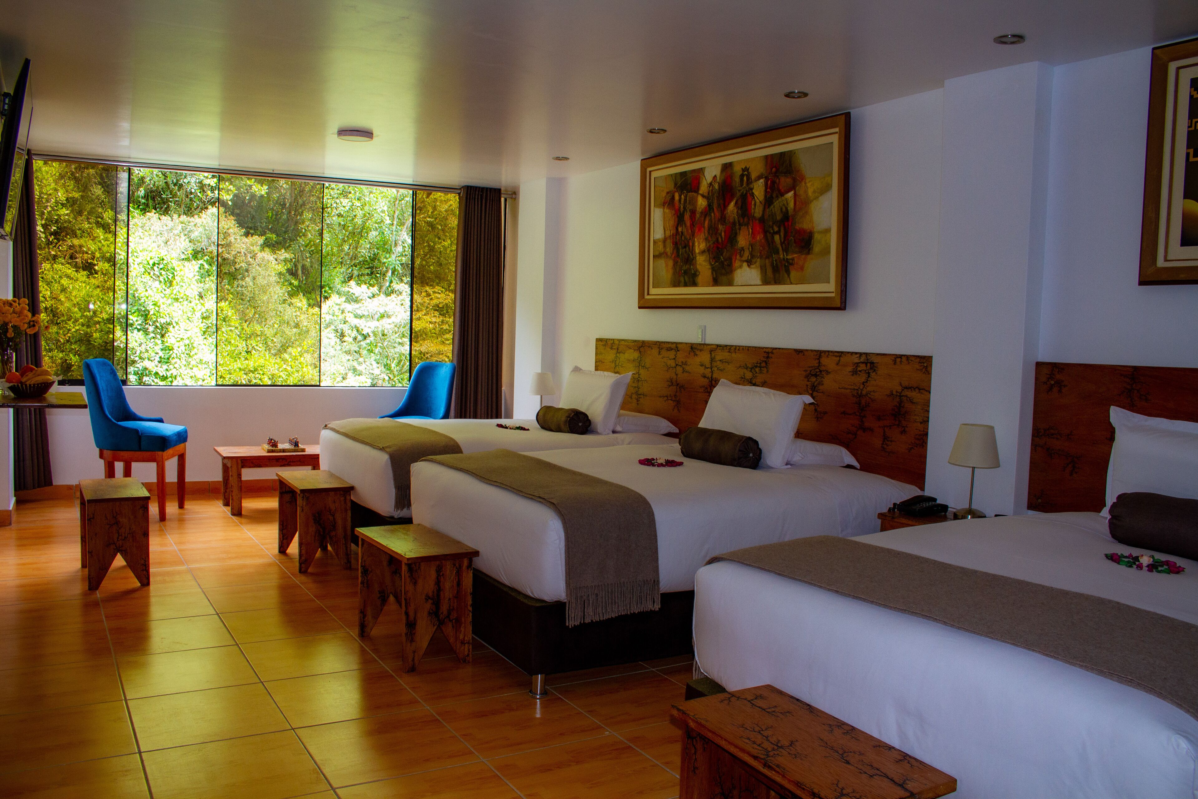 A hotel room with two beds, a desk, a chair, and a large window offering a view of greenery.