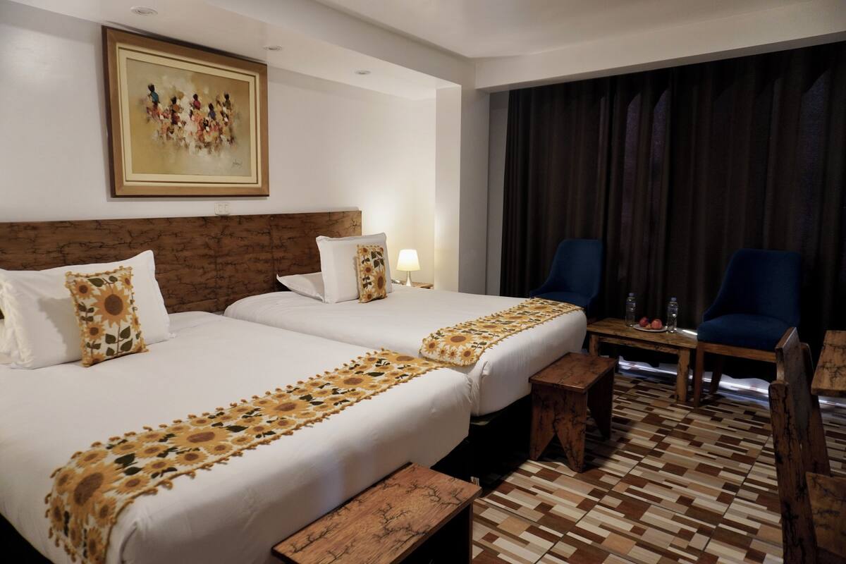 A hotel room with two beds, a wooden headboard, a painting on the wall, a desk with chairs, and a patterned floor.