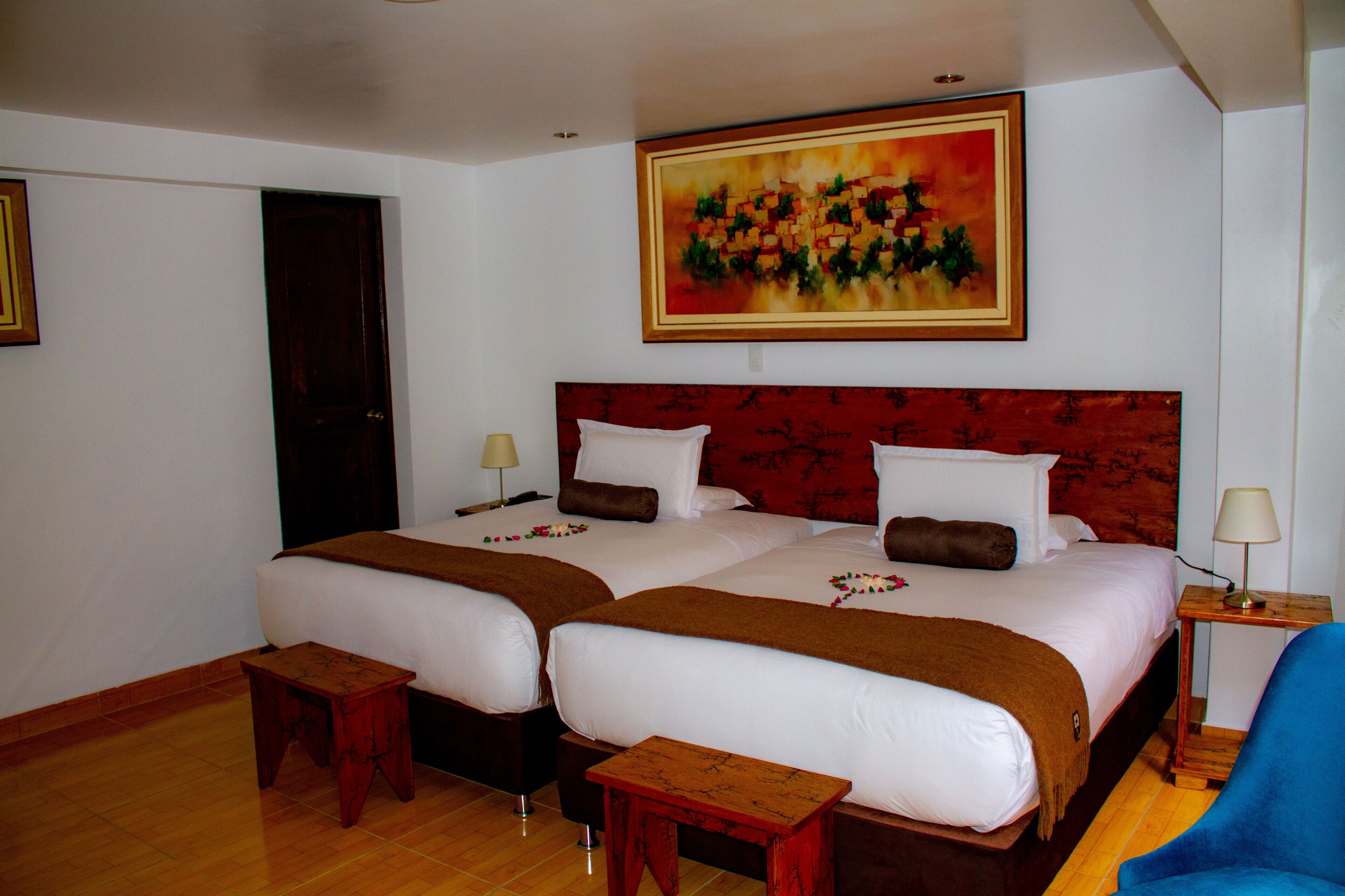 Habitación de hotel con dos camas, piso de madera, un cuadro sobre las camas y una silla azul.