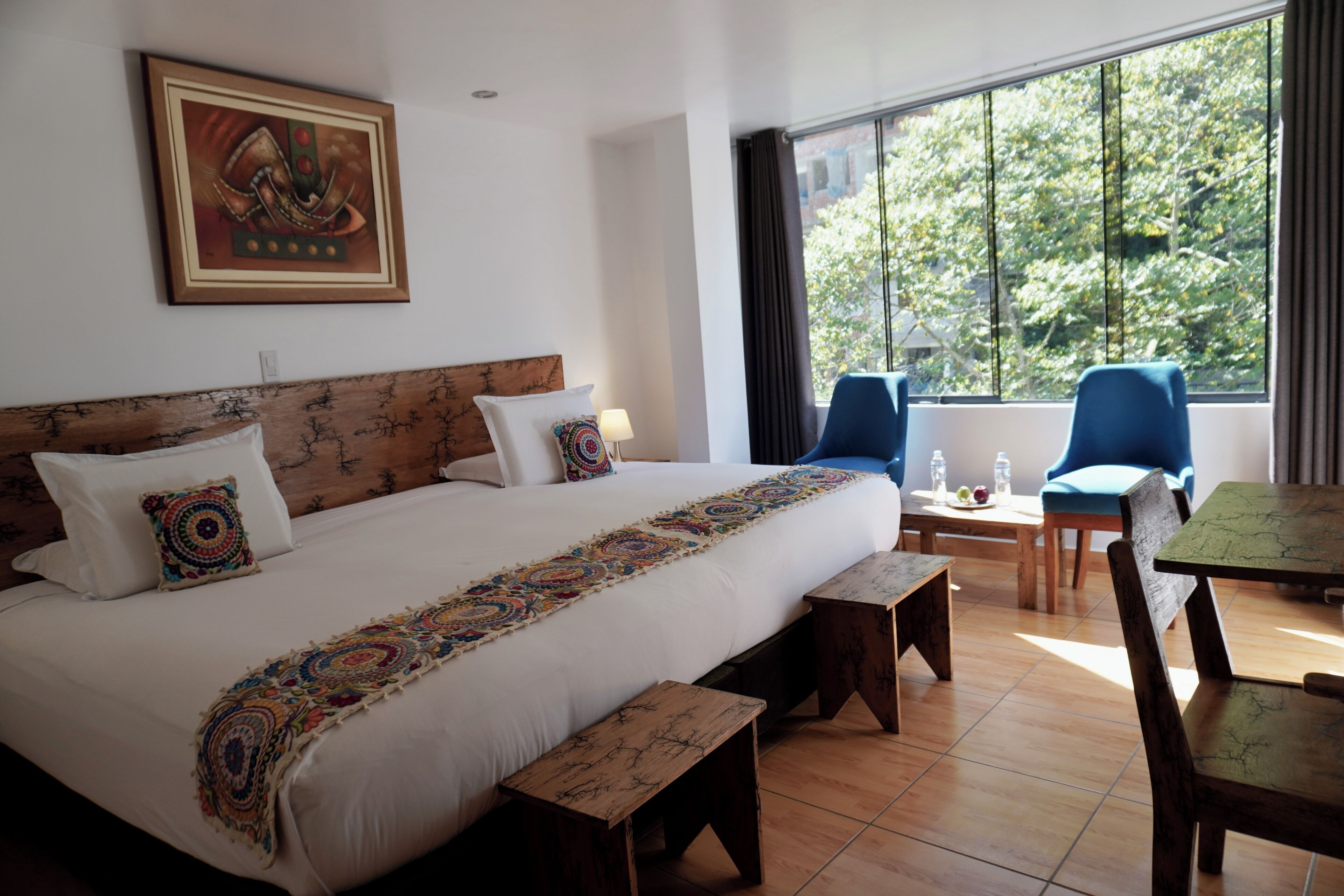 Quarto de hotel com uma cama grande, duas cadeiras, uma mesinha e uma janela com vista para árvores.