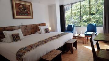 Quarto de hotel com uma cama grande, duas cadeiras, uma mesinha e uma janela com vista para árvores.