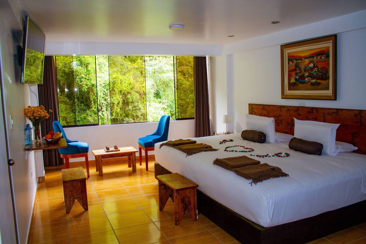 A hotel room with a large bed, two blue chairs, a wooden table, and a window with a view of trees.
