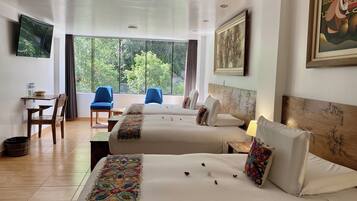 A hotel room with two beds, a desk, chairs, and a large window with a view of trees.