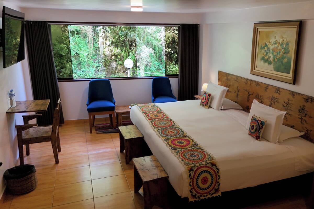 A hotel room with a large bed, a desk, two chairs, a window with a view of greenery, and a painting on the wall.
