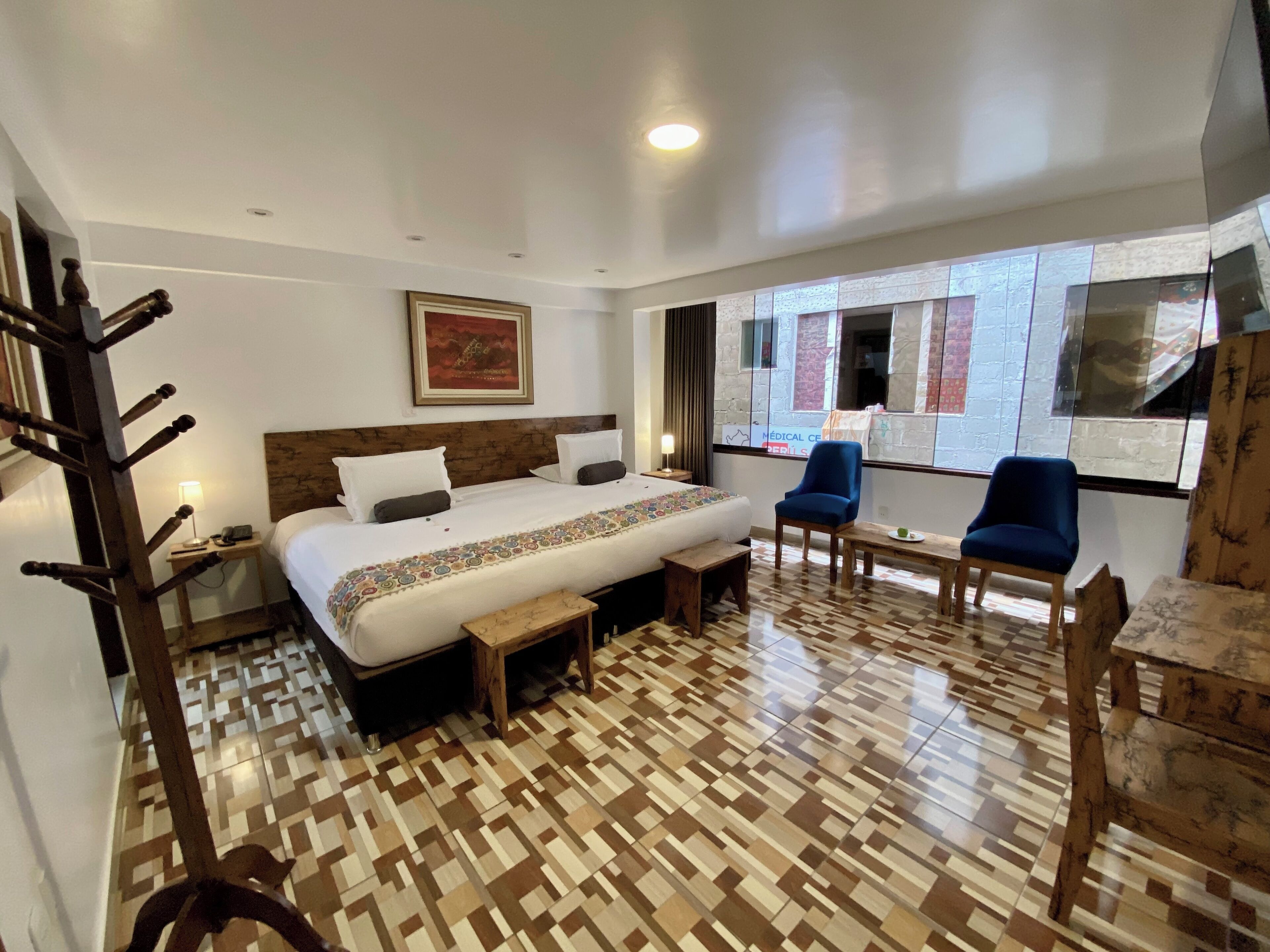 A hotel room with a large bed, a wooden bench, two blue chairs, a small table, and a window with a view of buildings.