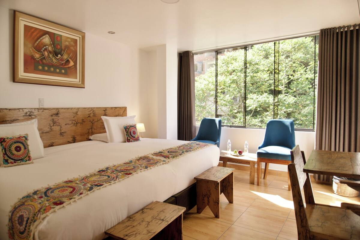 A hotel room with a large bed, a desk, two chairs, a window with curtains, and a painting on the wall.