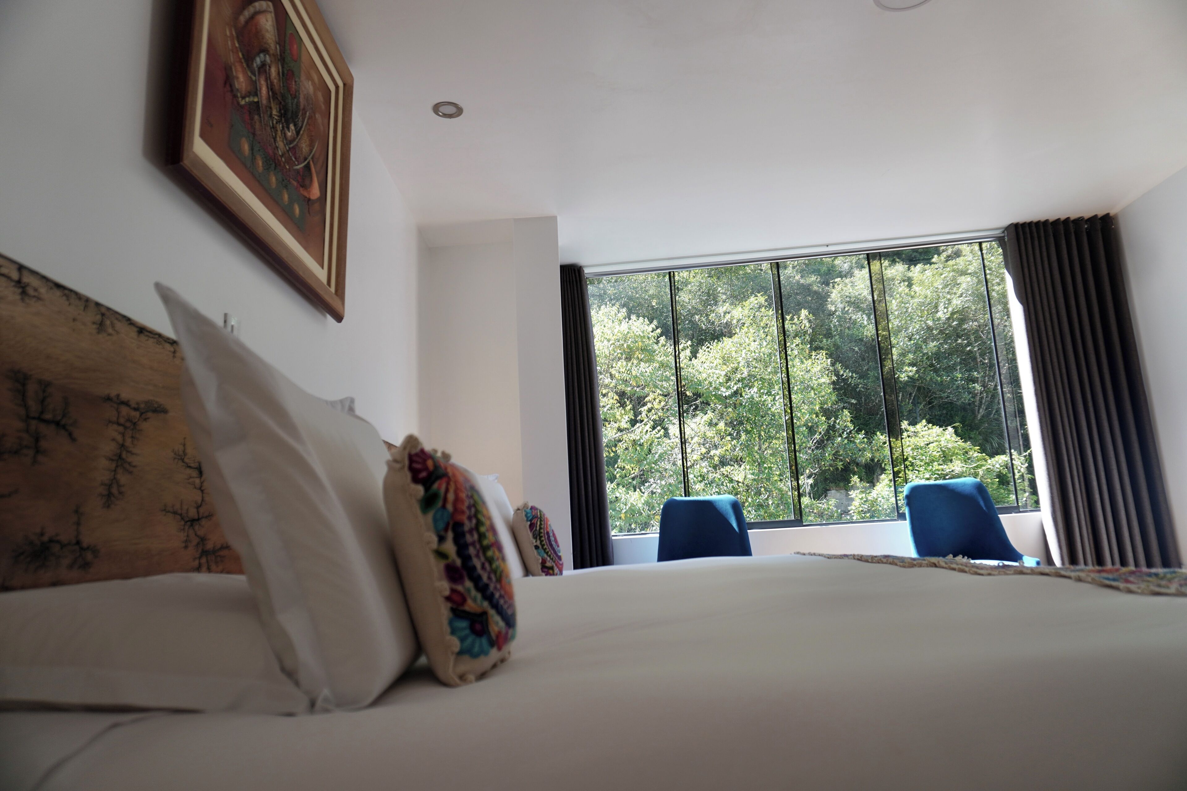 A hotel room with a large bed, a window with a view of trees, a blue chair, and a painting on the wall.
