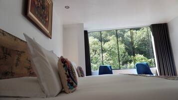 A hotel room with a large bed, a window with a view of trees, a blue chair, and a painting on the wall.