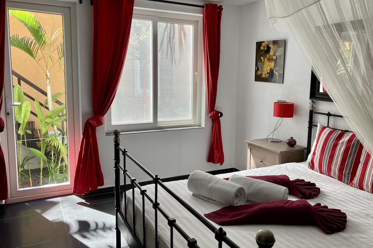 A hotel room with a bed, a nightstand, a lamp, and a window with red curtains.