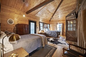 A cozy log cabin bedroom with a large bed, a smaller bed, a wooden wardrobe, a nightstand, a lamp, and a cowhide rug.