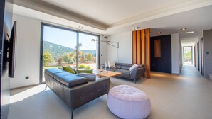 A modern living room with a sofa, ottoman, and coffee table, large windows, and a view of greenery. - Contemporary villa with pool and sea view for 10 people (Cavalaire-sur-Mer)
