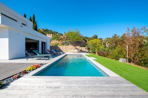 Un espace piscine moderne avec une terrasse en bois, des chaises longues et un ciel d’un bleu éclatant.