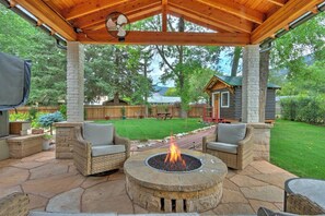 Un patio al aire libre con suelo de piedra, dos sillas de mimbre, un hogar de piedra y una pérgola de madera.