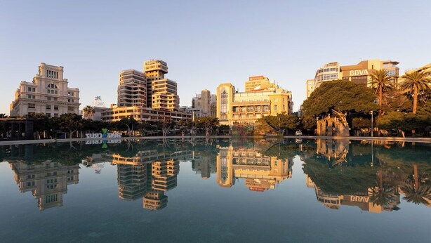 A cityscape with modern buildings reflected in a calm body of water.