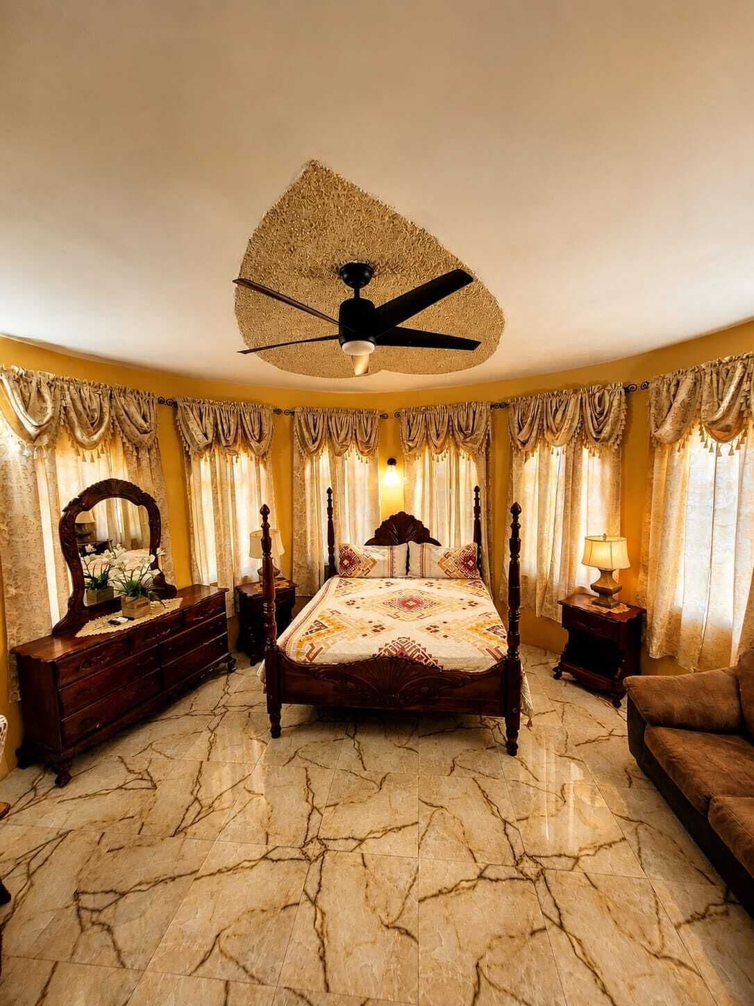 Une chambre à coucher comprenant un lit, une commode, un miroir, un ventilateur de plafond et des rideaux.