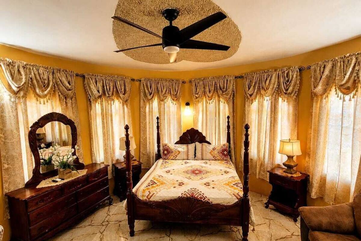 Une chambre à coucher comprenant un lit, une commode, un miroir, un ventilateur de plafond et des rideaux.
