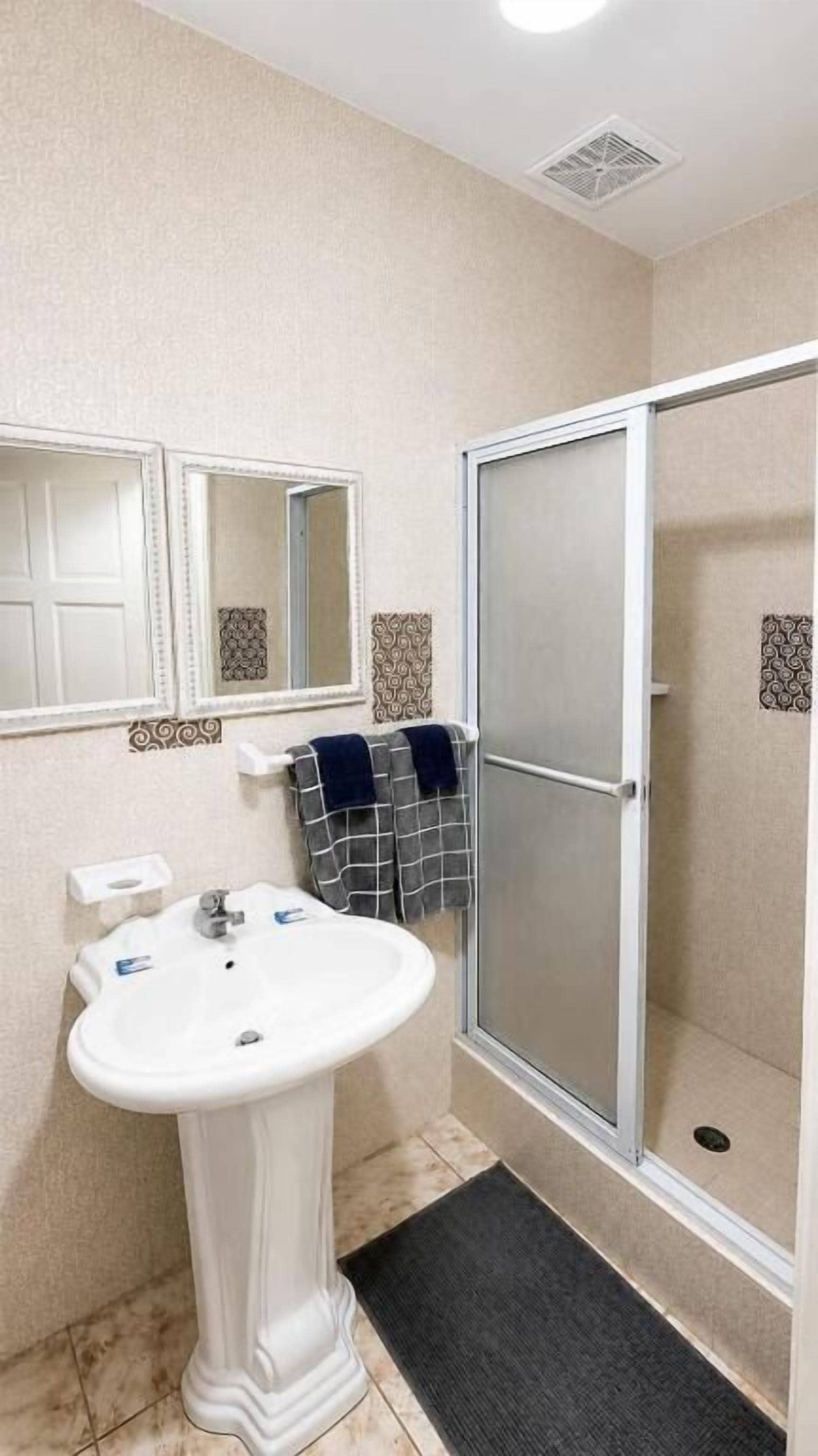 Une salle de bain avec un lavabo sur colonne, deux miroirs fixés au mur, un porte-serviettes avec des serviettes, une douche avec une porte en verre et des carreaux décoratifs sur les murs.