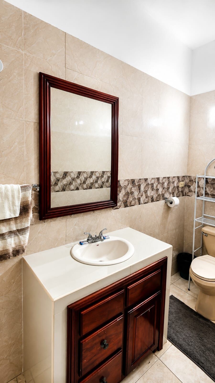 Een badkamer met een marmeren wand, een houten wastafelmeubel, een wastafel, een spiegel, een toilet en een handdoekhouder.