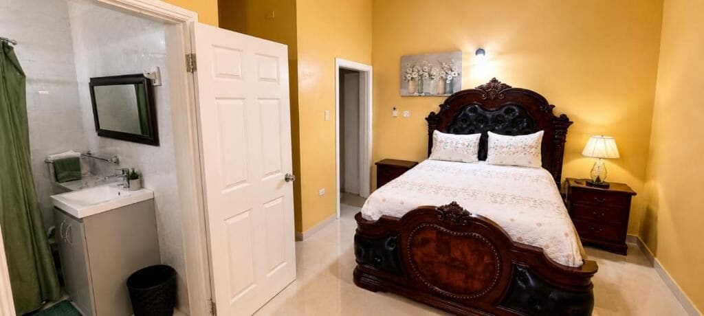 A bedroom with a wooden bed, a bedside table, a nightstand, a wall-mounted mirror, and a bathroom with a sink and shower curtain.