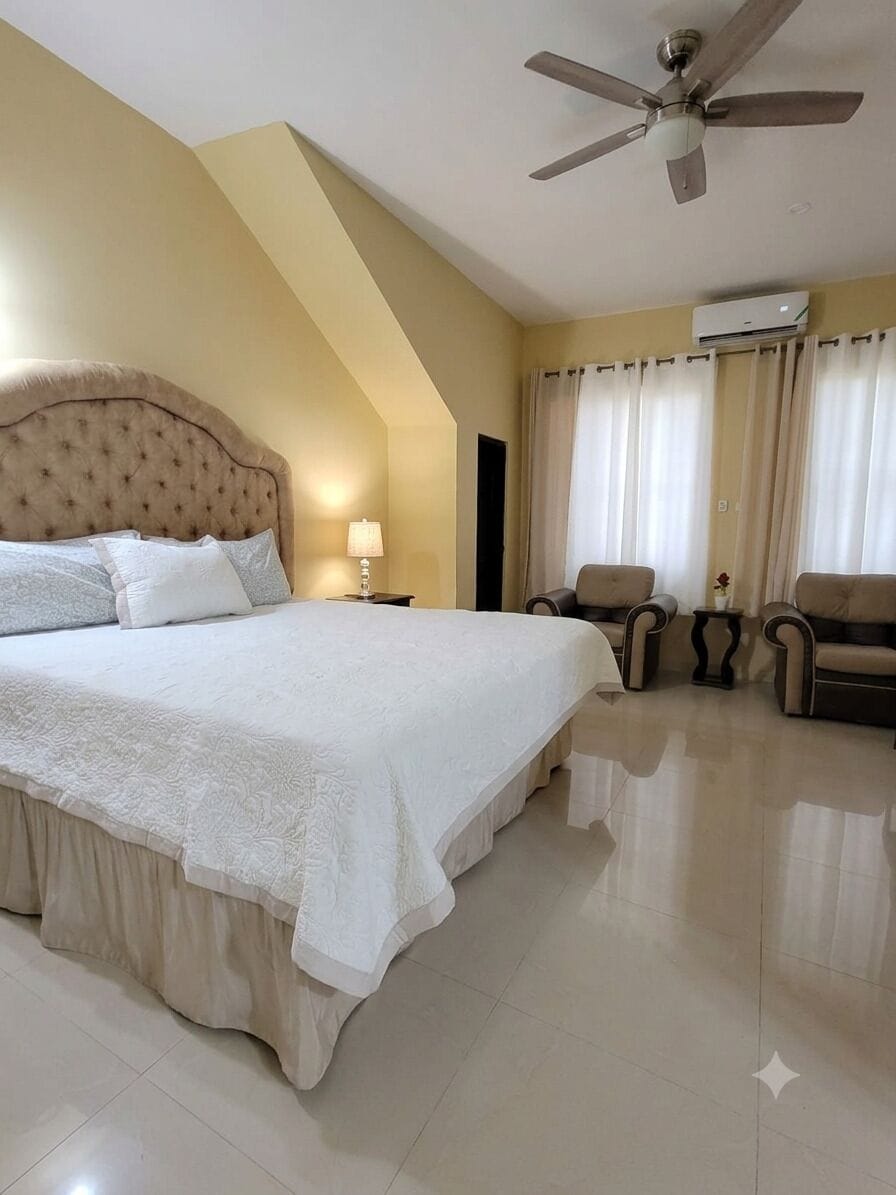 Une chambre d’hôtel avec un grand lit, un ventilateur de plafond, deux fauteuils, une petite table et un appareil de climatisation.