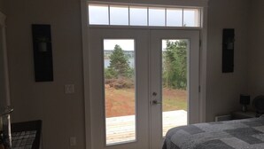 Quarto com uma grande porta de vidro deslizante que dá para um deck, cama com colcha cinzenta e uma pequena mesa com abajur.
