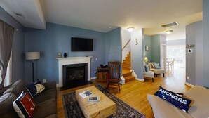A living room with a fireplace, a television mounted above it, a sofa with a cushion, a coffee table, and a staircase leading to another room. - Entire Townhouse in Rock Hall (Rock Hall)