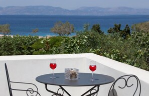 Ein Tisch mit zwei Gläsern Rotwein und einem dekorativen Glasgefäß, vor einer malerischen Kulisse aus Meer und Bergen.
