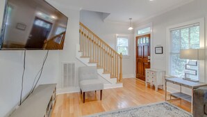 A spacious living area with a wooden staircase, a flat-screen TV mounted on the wall, a grey armchair, a small desk with a lamp, and a large window providing natural light. - Southern City Comfort - Huge Private Backyard (Nashville)