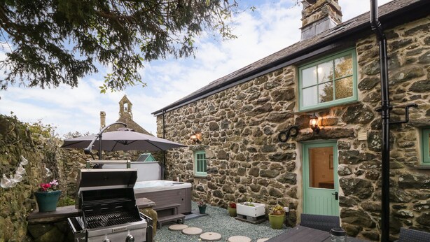 A stone building with a stone chimney, a BBQ grill, a hot tub, and outdoor seating. - Bryn Aber Bach (Pwllheli)