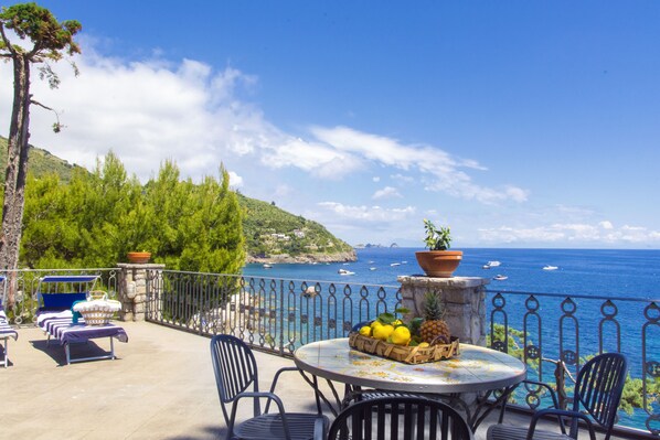 Une terrasse avec des sièges extérieurs, une table garnie de fruits et une vue sur la mer.