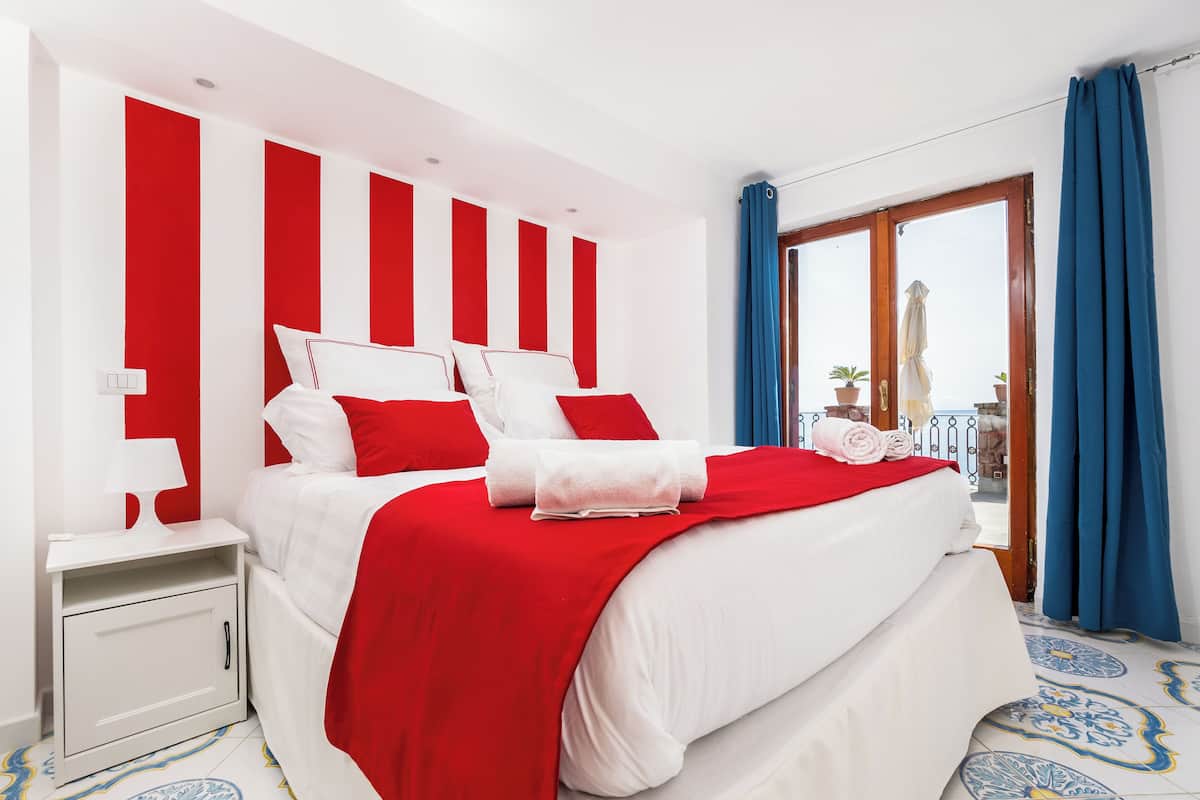 A hotel room with a large bed, white and red bedding, a nightstand, and a view of the outside balcony.
